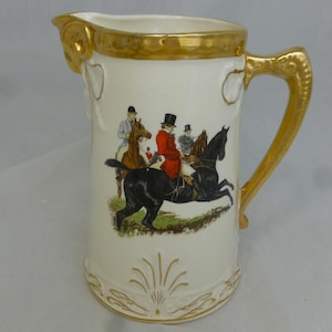 Peut inclure: Un pichet en céramique blanche avec un bord et une poignée dorés. Le pichet présente une scène peinte à la main de trois personnes à cheval dans un champ.