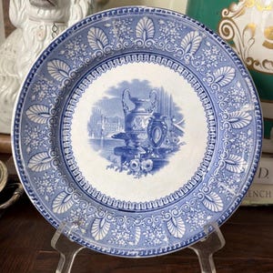 May include: A round ceramic plate featuring a blue and white transferware pattern. The center depicts a classical urn and vase scene, while the border is decorated with repeating floral motifs and shell-like shapes on a white background.