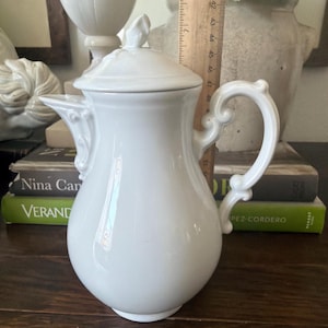 May include: A white ceramic coffee pot with a lid, featuring a curved handle and a spout. A wooden ruler is placed behind the pot, showing a height of approximately 9 inches from the base to the top of the lid finial. The pot has a smooth finish.