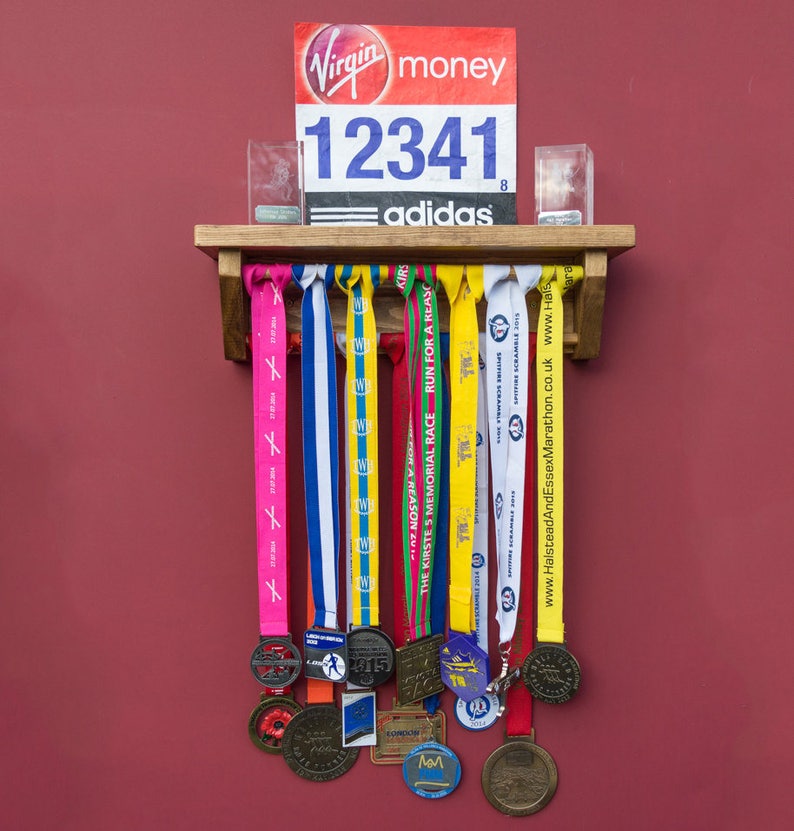 May include: A wooden shelf with a collection of colorful medals hanging from it. The medals are from various races and events, including the "Kirstes Memorial Race Run for a Reason" and the "Spitfire Scramble". The shelf also has a number "12341" and the words "Virgin money" and "adidas" on it.