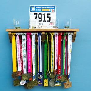 May include: A wooden medal display rack with a variety of colorful ribbons and medals hanging from it. The rack has a shelf on top with a race bib that reads "7915 NEIL".