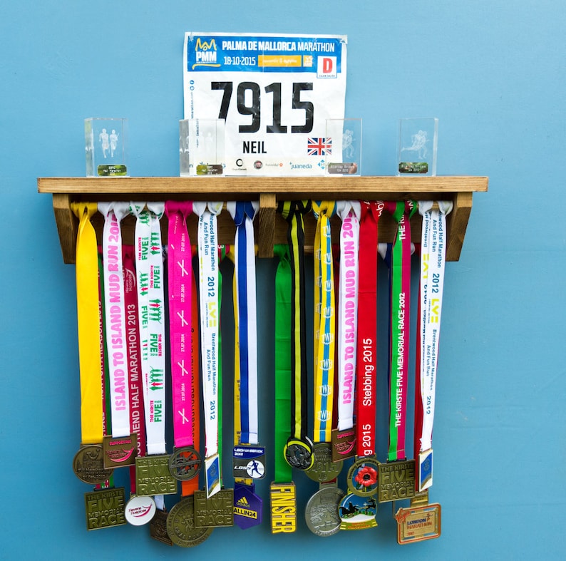 May include: A wooden medal display rack with a variety of colorful ribbons and medals hanging from it. The rack has a shelf on top with a race bib that reads "7915 NEIL".