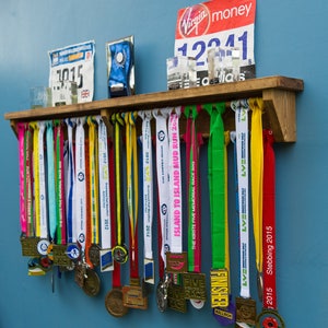 May include: A wooden shelf with a collection of colorful medals hanging on hooks. The medals are from various races and events, including the "Island to Island Mud Run" and the "Kirst Five Memorial Race".