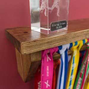 May include: A wooden shelf with a clear glass trophy on top. The trophy has a silver plaque that reads "Billericay Striders 10k 2010". The shelf is decorated with colorful ribbons.