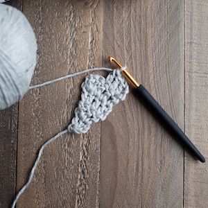 May include: A close-up of a gray crochet project in progress. A black crochet hook is being used to work the stitches. Two balls of gray yarn with the label "Wool and the Gang" are in the background.
