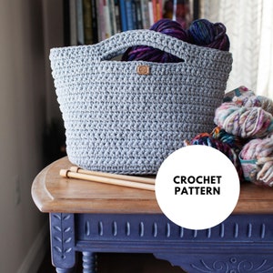 Puede incluir: Una bolsa de mano de crochet gris con asas de madera, sentada sobre una mesa de madera con ovillos de lana y agujas de tejer. El texto "CROCHET PATTERN" está en un círculo blanco.