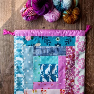 May include: A drawstring project bag featuring a patchwork design with various fabric patterns including greyhounds, flowers, and shoes, paired with four skeins of multi-colored yarn in shades of purple, blue, and brown.