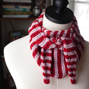 May include: A red and white striped knitted cowl scarf displayed on a mannequin. The scarf is made of thick yarn and features alternating stripes of red and white. The cowl is designed to be worn around the neck and has a cozy, handmade appearance.