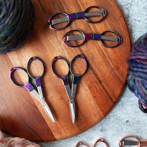 May include: A wooden cutting board with five pairs of scissors. Two pairs are iridescent with silver blades, and three pairs are rose gold with silver blades. Two balls of yarn are in the background.