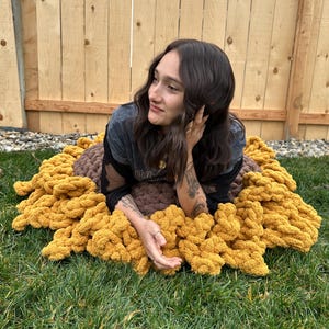 Puede incluir: Una gran manta de crochet de color amarillo mostaza y marrón con un borde de flecos. La manta está colocada sobre un lecho de hierba verde.