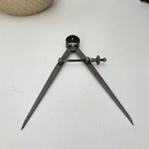 May include: A vintage metal drafting compass with a dark grey finish, featuring two pointed legs connected by a central pivot point and an adjustable threaded screw mechanism for precise measurement and drawing of circles and arcs.