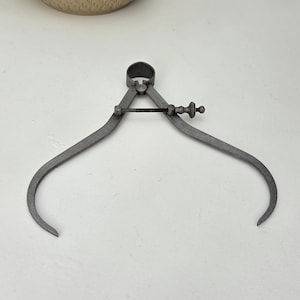 Peut inclure: Un outil de mesure de type compas d'épaisseur en métal gris mat, doté de deux branches courbes reliées par un pivot central et d'une tige filetée réglable munie d'un petit bouton pour ajuster l'écartement avec précision.