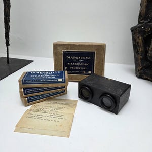 May include: A vintage stereoscope with two lenses, alongside boxes of glass slides marked "DIAPOSITIVE SU VETRO STEREOSCOPIO PROIEZIONI" and a pile of old papers. The stereoscope is black in colour.