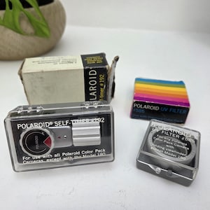 Op de afbeelding: Een Polaroid zelfontspanner #192 in een doorzichtige plastic behuizing met de originele doos, naast een Polaroid UV-filter #585 in een doorzichtige behuizing met een doos met regenboogstrepen. De zelfontspanner is zilver en zwart van kleur.