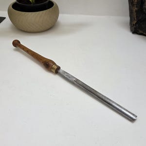May include: A woodturning gouge featuring a long, slender steel blade with a concave profile and a rounded tip. The tool has a polished wooden handle with a prominent grain pattern, connected to the metal shaft by a brass ferrule.