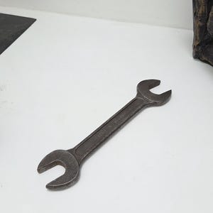 Puede incluir: Una llave inglesa gris oscura, probablemente de acero, descansa sobre una superficie blanca. La llave tiene dos extremos abiertos de diferentes tamaños. El texto "MADE IN POLAND" está estampado en el mango. Esta herramienta se utiliza para apretar o aflojar tuercas y pernos.