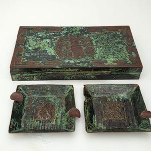 Pode incluir: Um conjunto de itens de bronze antigos, incluindo uma caixa retangular e duas cinzeiros quadrados. A caixa e os cinzeiros têm uma pátina verde com detalhes em marrom e desenhos em relevo. Os cinzeiros têm um pequeno descanso arredondado para cigarros.