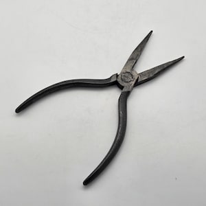 May include: A pair of dark grey metal needle-nose pliers with long, tapered jaws and curved handles. The central pivot point features a circular stamp with the text "SAFETY" visible on the metal surface. The tool has a weathered, matte finish.