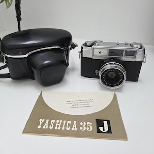 May include: A vintage Yashica 35 J camera with a black leather case and instruction booklet. The camera is black and silver with a lens that reads 'Yashinon 1:2 f=45mm'. The booklet reads 'Yashica 35 J'.
