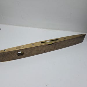 Puede incluir: Un nivel de madera vintage con detalles de latón. El nivel tiene forma rectangular con una parte superior de latón y un agujero circular. El texto del nivel es parcialmente visible. Una herramienta clásica para carpintería y construcción.