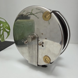 Puede incluir: Un carrete de pesca vintage, de forma circular, con un cuerpo de metal plateado pulido. El carrete presenta detalles en color latón, un asa y un pequeño botón de control. El diámetro es de aproximadamente 10 cm.