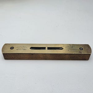 May include: A rectangular spirit level featuring a brass top plate and a dark wooden base. The brass plate is engraved with the text TRADE MARK HOCKLEY ABBEY and includes a central glass vial. Two dark screws secure the brass plate to the wood.