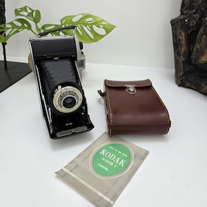 May include: A vintage Kodak Junior I folding camera with a black bellows, a brown leather carrying case with a metal clasp, and an instructional booklet titled How to Use Your Kodak Junior I Camera, Kodak Limited London, printed on a green background.