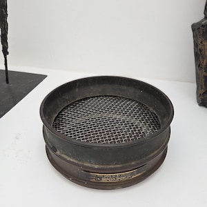 May include: A circular metal sieve with a fine wire mesh. The sieve has a dark, aged appearance with a wide rim. The mesh is silver, and the metal frame is dark. It is likely used for sifting dry ingredients, such as flour or sugar.