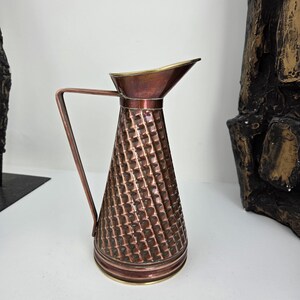 May include: A conical copper pitcher featuring a textured diamond-patterned body, a smooth flared rim, and a thin angular handle. The metal surface displays a warm reddish-brown tone with polished brass accents along the base and the top edge.