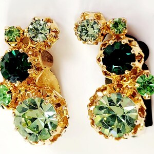 May include: A pair of gold-toned clip-on earrings featuring clusters of green and emerald-colored rhinestones. The earrings have a vintage aesthetic, with intricate metalwork surrounding the stones. The design includes various sizes and shades of green.