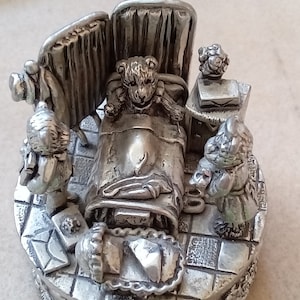Könnte beinhalten: Eine silberne Zinnfigur, die eine Szene mit einem Teddybären in einem Krankenhausbett zeigt, den zwei andere Teddybären besuchen. Die Figur steht auf einem runden Sockel mit einem karierten Muster.