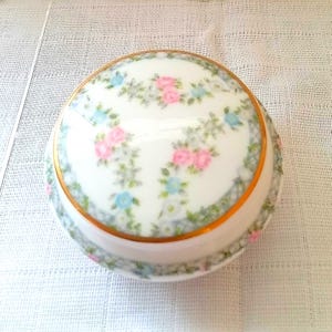 May include: A round white porcelain trinket box with a lid featuring a delicate floral pattern of pink roses and small blue flowers. The lid is bordered by a thin gold rim, and the base of the container is decorated with a matching floral garland.