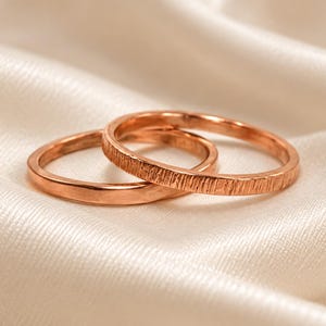 May include: Two rose gold rings, one with a smooth, polished finish and the other featuring a textured, vertical line pattern. The rings are circular bands crafted from a metallic material with a warm, reddish-pink hue, displayed together.