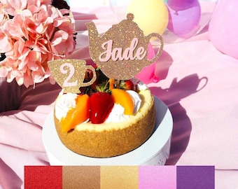 May include: A gold glitter teapot and teacup cake topper with the name "Jade" written on the teapot. The topper is on a cheesecake with fruit on top. The background is pink with a clear balloon with smaller balloons inside.