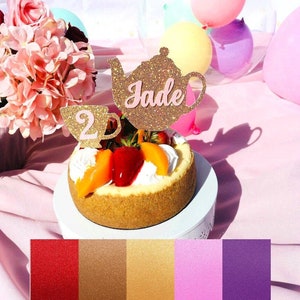 May include: A gold glitter teapot and teacup cake topper with the name "Jade" written on the teapot. The topper is on a cheesecake with fruit on top. The background is pink with a clear balloon with smaller balloons inside.
