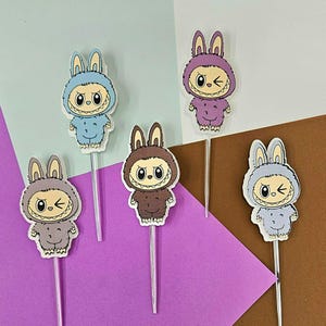 May include: A collection of whimsical cake toppers featuring cartoon characters in bunny costumes. The toppers are in various colors, including blue, purple, and brown, with clear plastic stems. Each character has a unique expression.