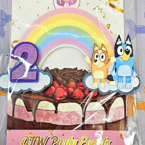 Puede incluir: Un conjunto de adornos para tartas con un arco&iacute;ris, dos perros de dibujos animados y un n&uacute;mero 2 morado. Los perros son del programa Bluey. El adorno est&aacute; sobre una tarta rosa y marr&oacute;n con chocolate y frambuesas. El texto "OTW Party Events" est&aacute; en la parte inferior de la imagen.