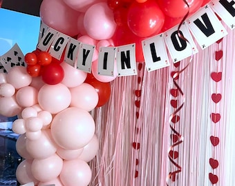 May include: A pink and red balloon arch with a banner that reads "Lucky in Love". The balloons are arranged in a cascading pattern and are decorated with playing cards. The background is a pink and white streamer curtain.
