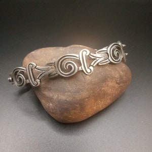 May include: A silver bracelet with a repeating pattern of stylized swirls. The bracelet is resting on a brown rock.