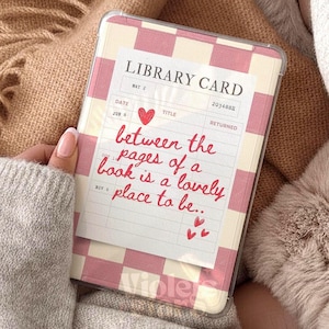 Puede incluir: Una funda transparente para teléfono con un diseño a cuadros rosa y blanco, con un gráfico de tarjeta de biblioteca. La tarjeta tiene el texto "between the pages of a book is a lovely place to be..." en escritura roja, con un corazón rojo.