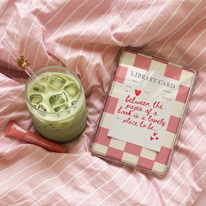 May include: A glass of matcha latte with ice cubes and a straw with a teddy bear charm. A pink lip product and a phone case with a pink and white checkered pattern and the text "Library Card" are also visible on a pink striped bedsheet.