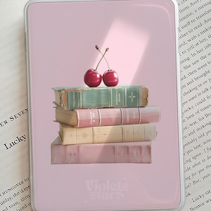 Op de afbeelding: Roze telefoonhoesje met een stapel vintage boeken in pastelkleuren en twee kersen. De tekst "Violets stars" staat onderaan. Op de achtergrond een pagina uit een boek met het woord "Lucky".