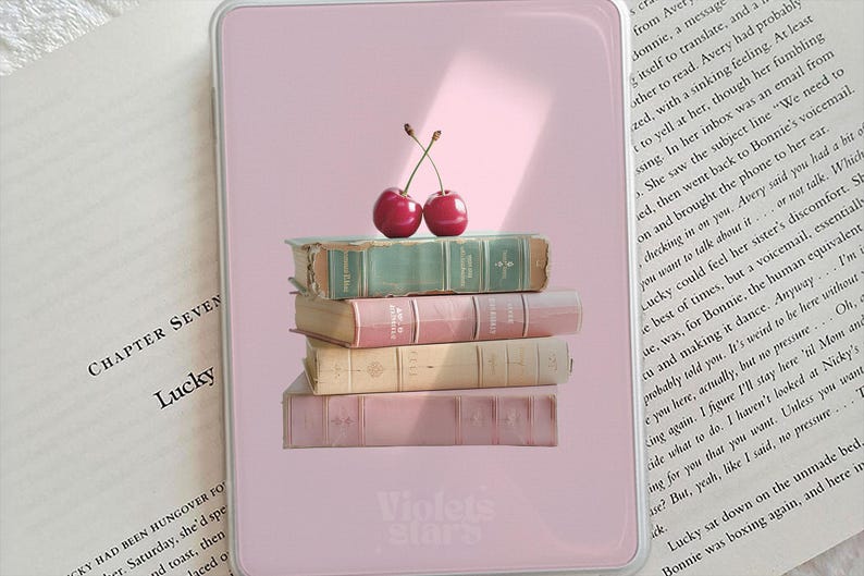 Pu&ograve; includere: Una custodia rettangolare per e-reader color rosa chiaro con una grafica di quattro libri impilati in tonalit&agrave; verde acqua, rosa, beige e rosa chiaro, sormontati da due ciliegie rosse. La parte inferiore della custodia riporta il testo Violet&eacute; stars.