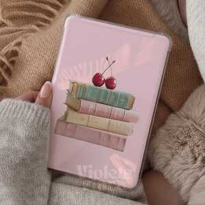 Pu&ograve; includere: Una custodia rettangolare per tablet con sfondo rosa chiaro che presenta una pila di quattro libri nelle tonalit&agrave; verde, rosa, crema e malva, sormontata da due ciliegie rosse. La parola Violets &egrave; stampata in bianco nella parte inferiore.