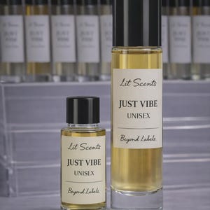 May include: Two glass perfume bottles with black caps and white labels that read "Lit Scents JUST VIBE UNISEX Beyond Labels." The bottles are filled with a light amber liquid. A row of similar bottles is in the background.