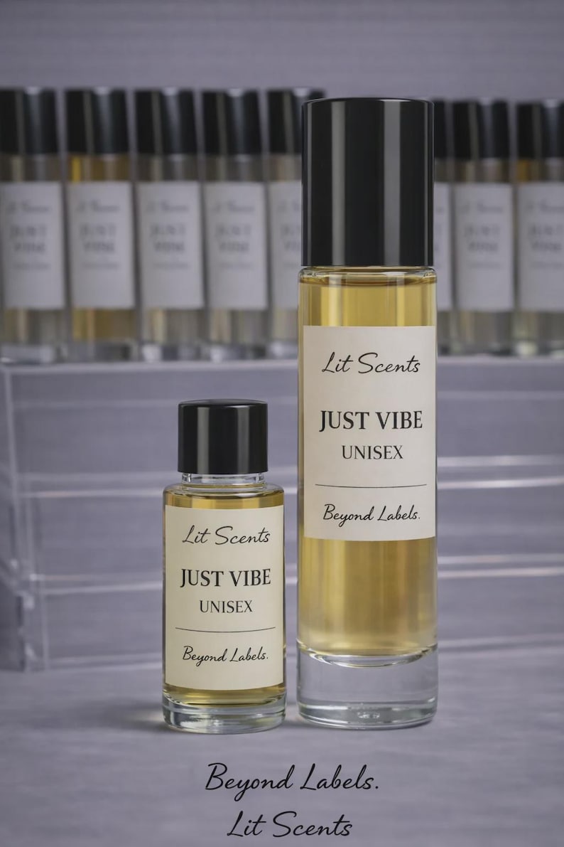 May include: Two glass perfume bottles with black caps and white labels that read "Lit Scents JUST VIBE UNISEX Beyond Labels." The bottles are filled with a light amber liquid. A row of similar bottles is in the background.