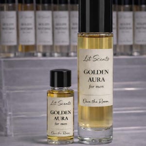 May include: Two glass perfume bottles with black caps, one large and one small, filled with a golden liquid. The labels read "Lit Scents GOLDEN AURA for men." The phrase "Own the Room" is also visible. Other bottles are in the background.
