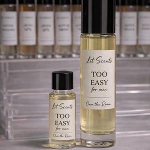 May include: Two glass perfume bottles with black caps, one large and one small, filled with a light yellow liquid. The bottles have white labels with the text "Lit Scents TOO EASY for men" and "Own the Room."