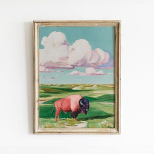 May include: Framed art print featuring a bison in a field. The bison is painted in shades of pink and purple, with a dark blue head. The background includes a green landscape, blue sky, and fluffy white clouds. The frame is a light gold color.