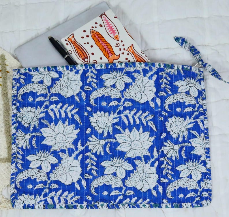 May include: A rectangular quilted fabric sleeve featuring a vibrant blue background with a repeating white floral and leaf pattern. The sleeve has a matching blue fabric tie closure on the right side and contains a white notebook with orange fish.